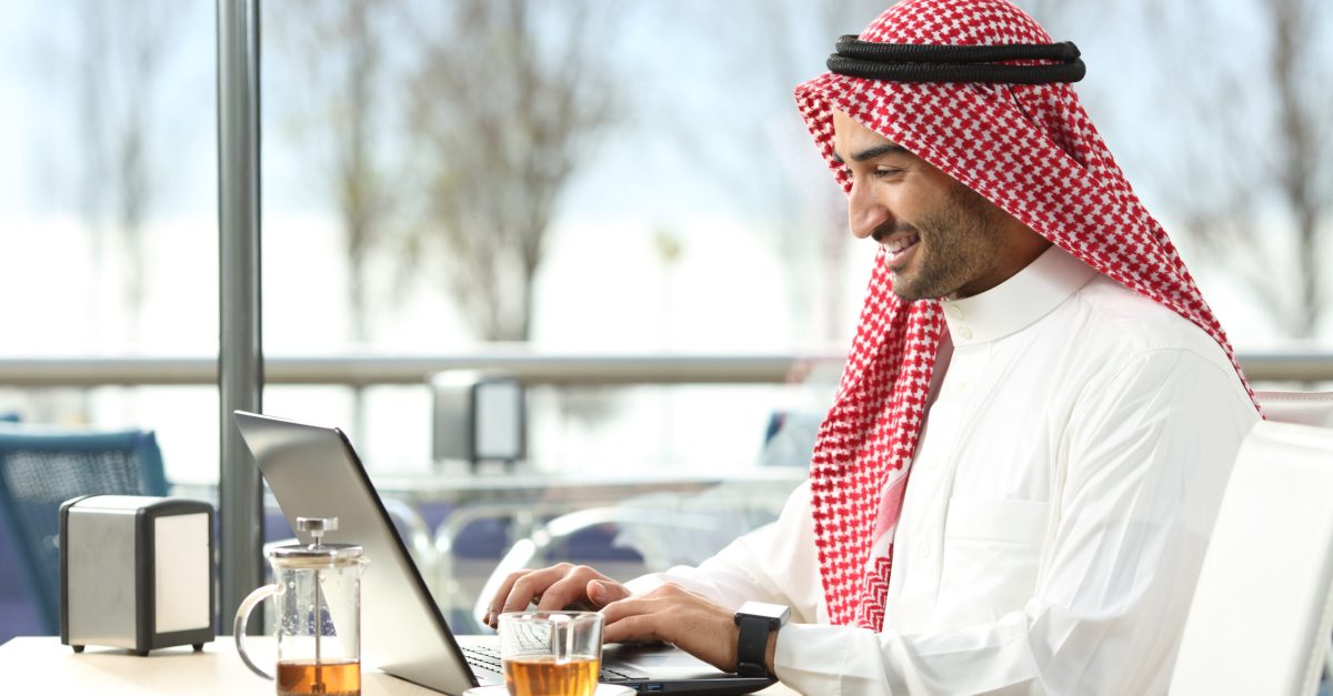 Arab saudi man working online with a laptop and smartwatch in a coffee shop or an hotel bar with a window and outdoor terrace in the background